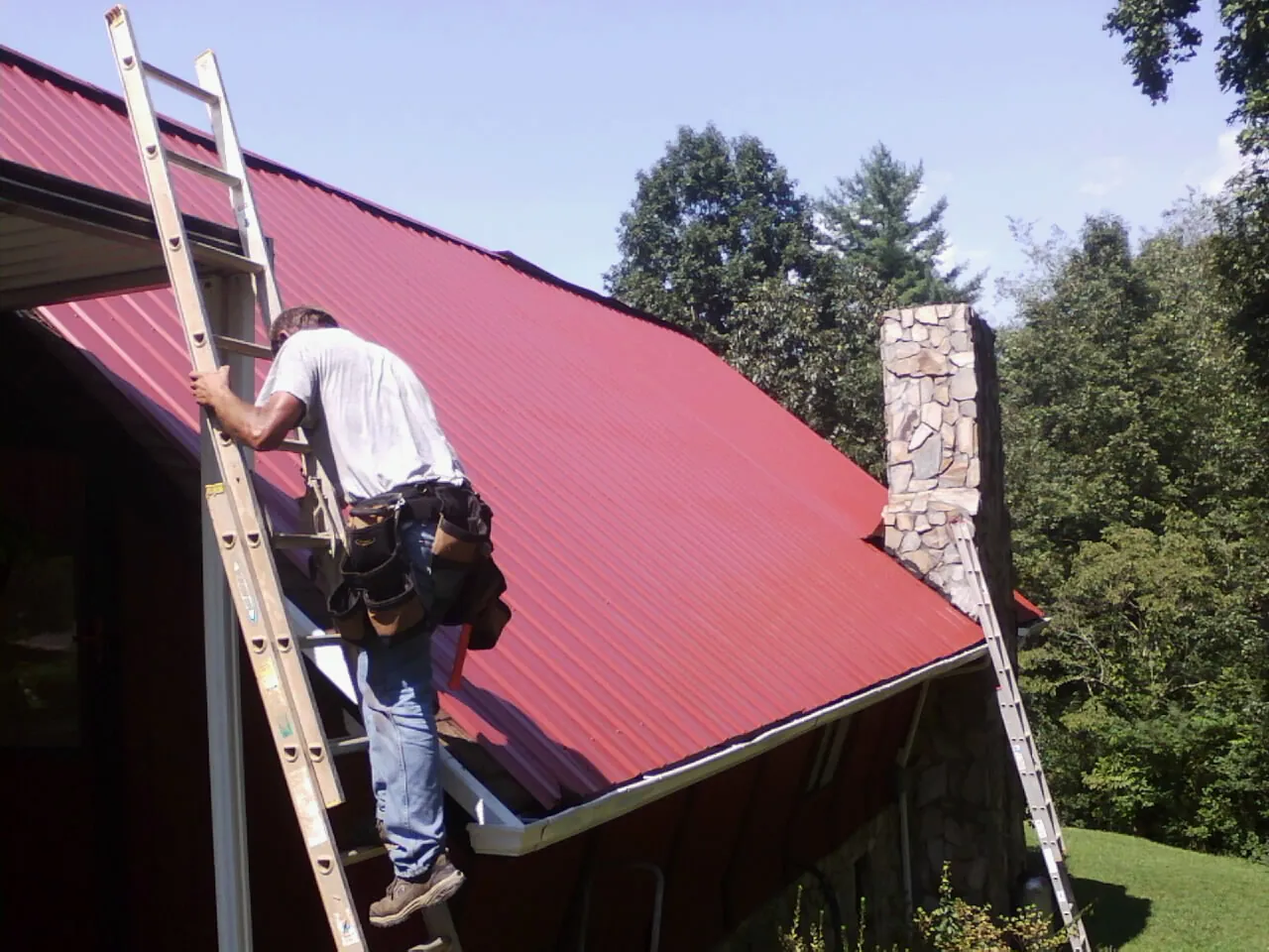 Roofer on a ladder beside a red metal roof and stone chimney — Soffit & Fascia Repair by Roofing Brothers in Eagle River
